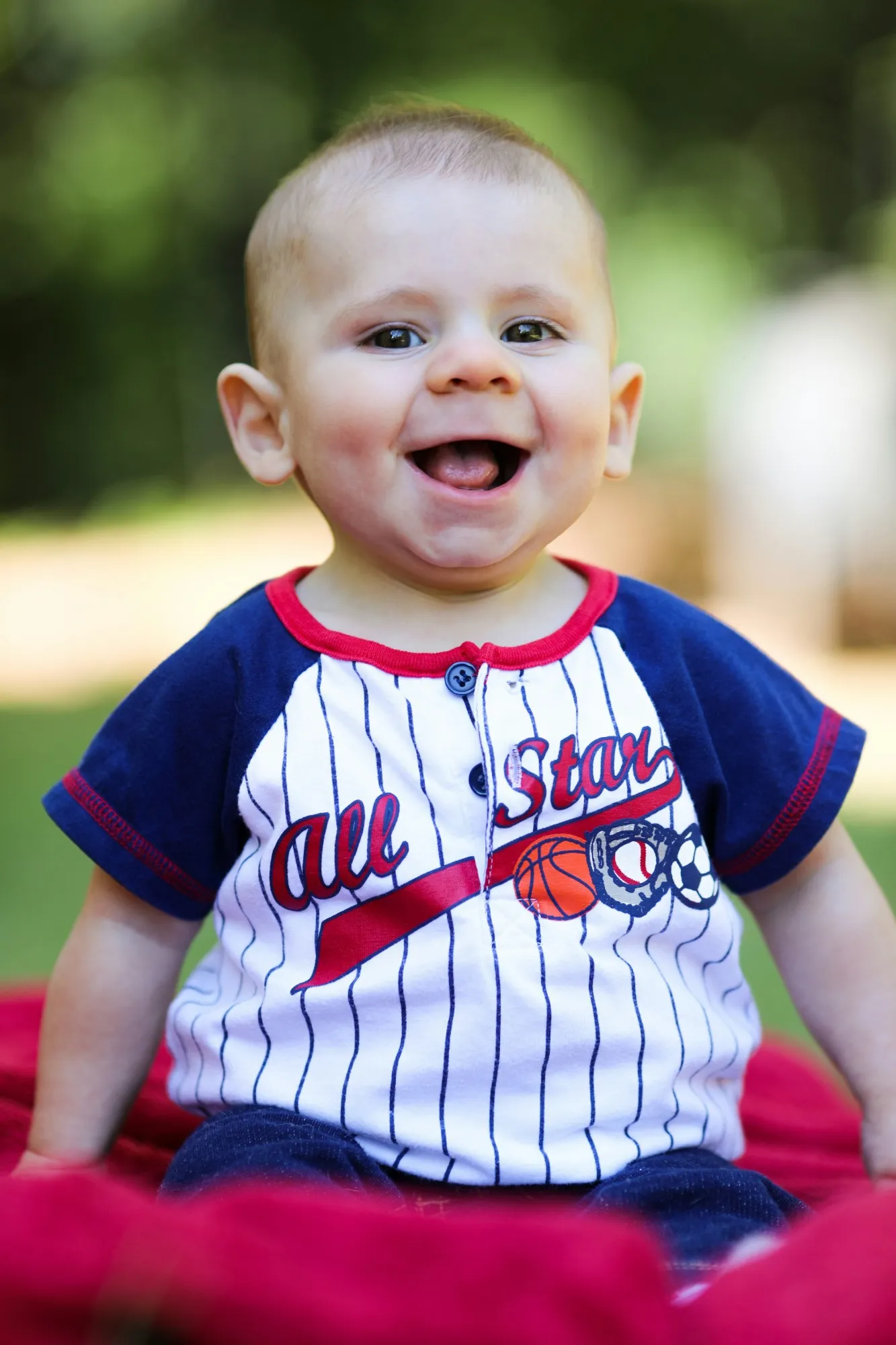 Laughing baby in a striped All Star shirt sitting on a red blanket outside