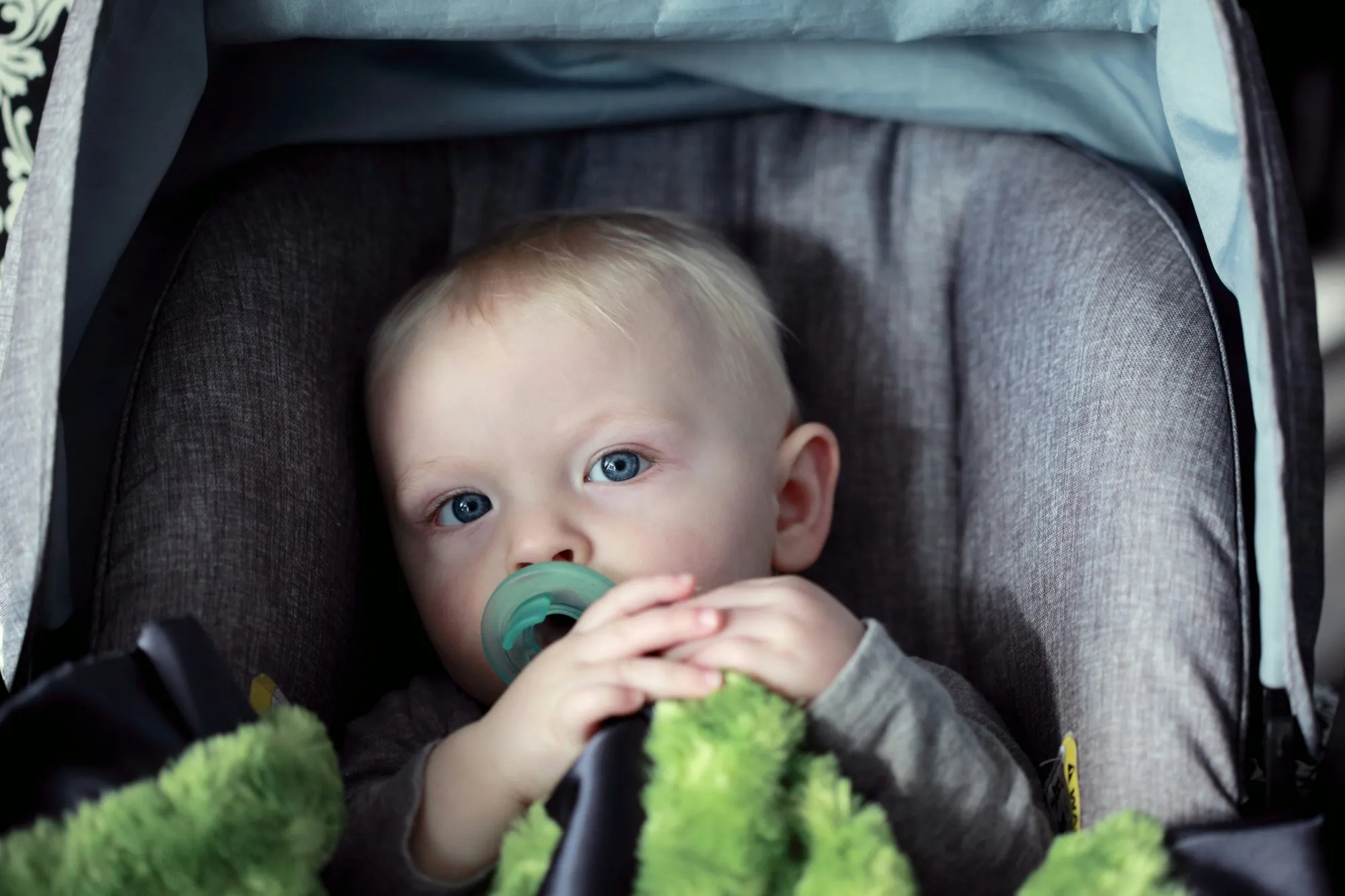 Baby with a pacifier and a green blanket, sitting in a gray infant car seat and looking up