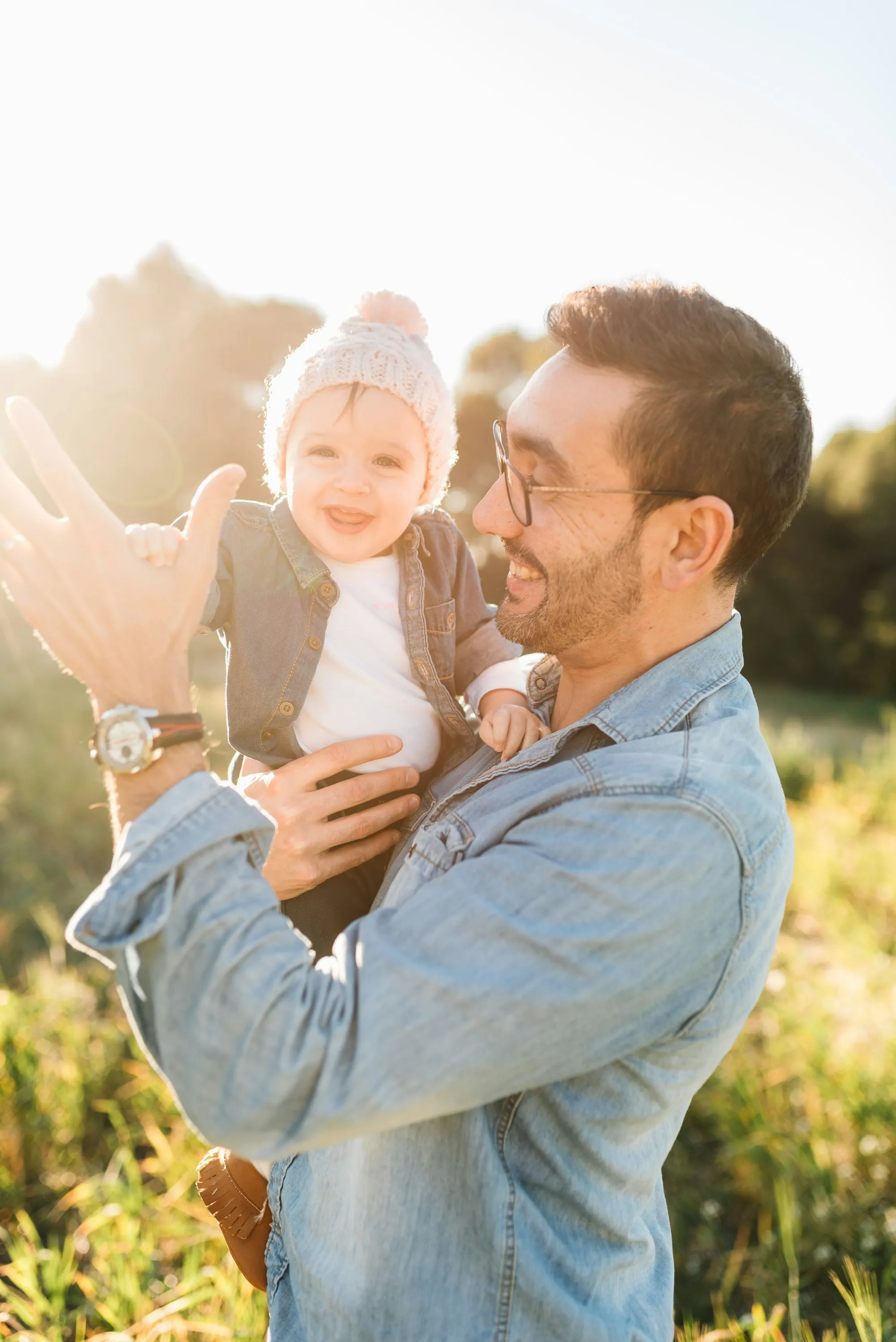 Smiling dad in a denim shirt holding his baby in a sunny meadow