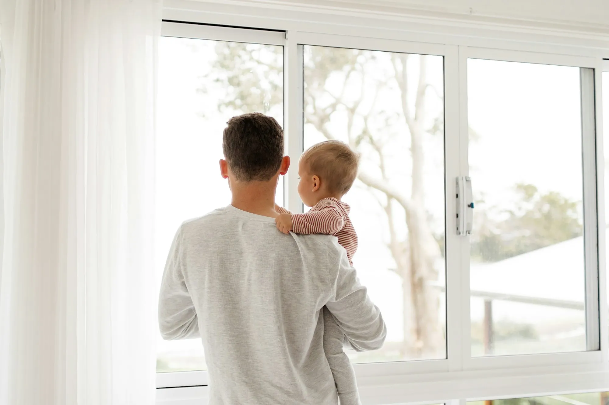 Dad holding his baby on his arm, both looking out of a large window
