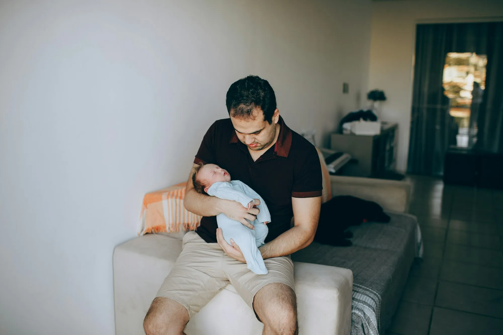 Dad sitting on the edge of a sofa cradling his newborn in his arms