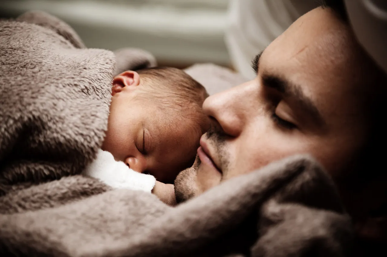 Dad resting with his eyes closed, his sleeping newborn tucked against his cheek under a soft brown blanket