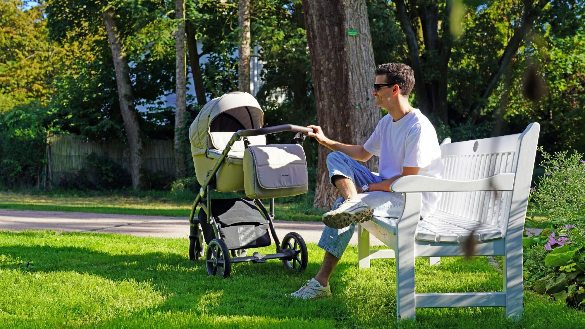 Smiling dad in sunglasses on a white park bench, one hand on the handle of a stroller on the grass