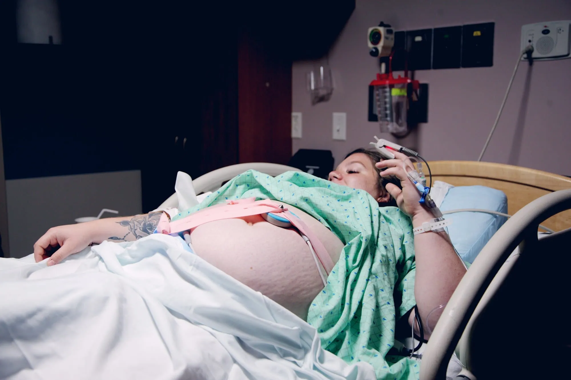 Pregnant woman lying in a hospital bed with monitor belts around her belly and an IV in her hand