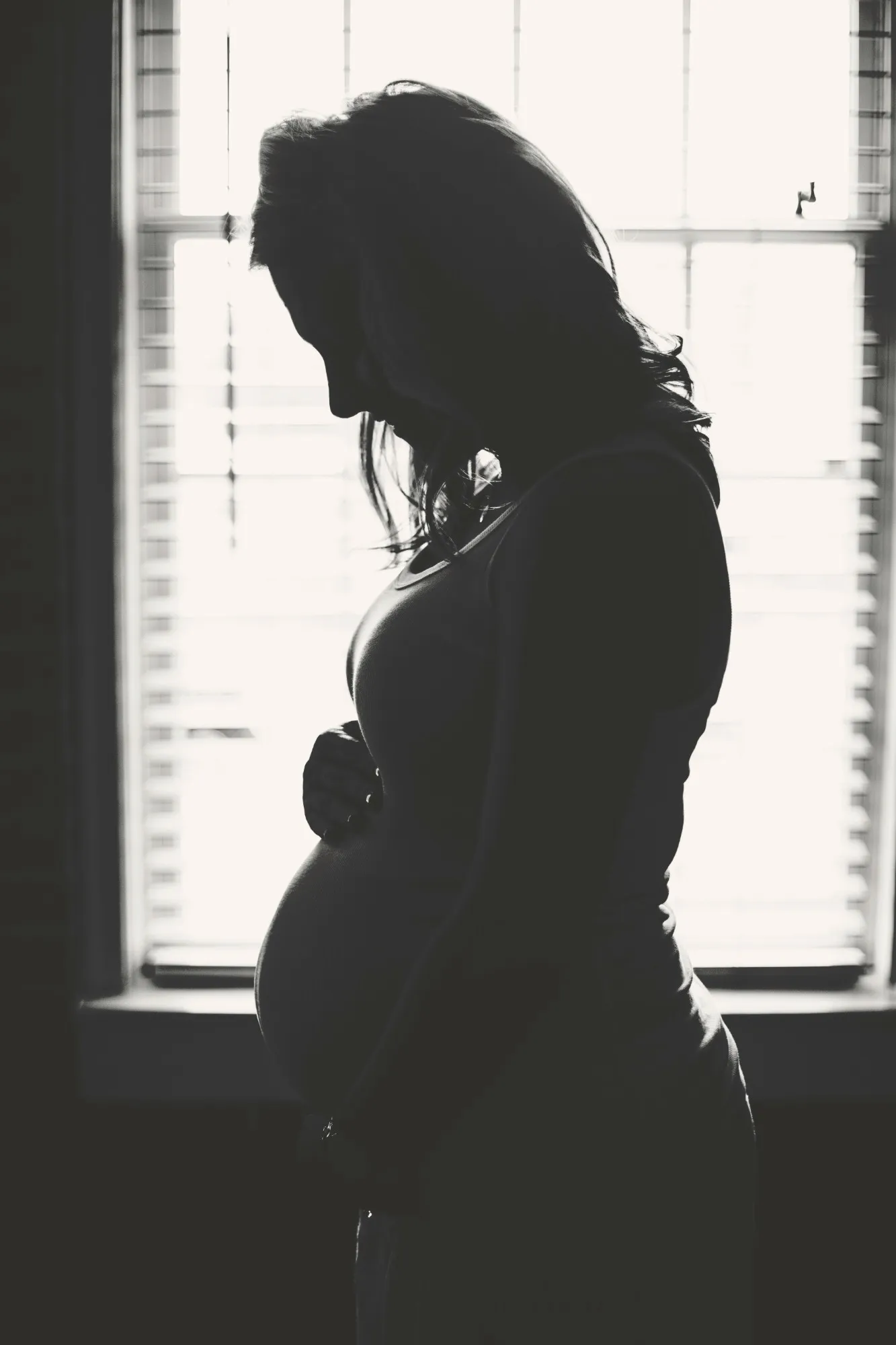 Pregnant woman in silhouette by a bright window, looking down with one hand on her belly